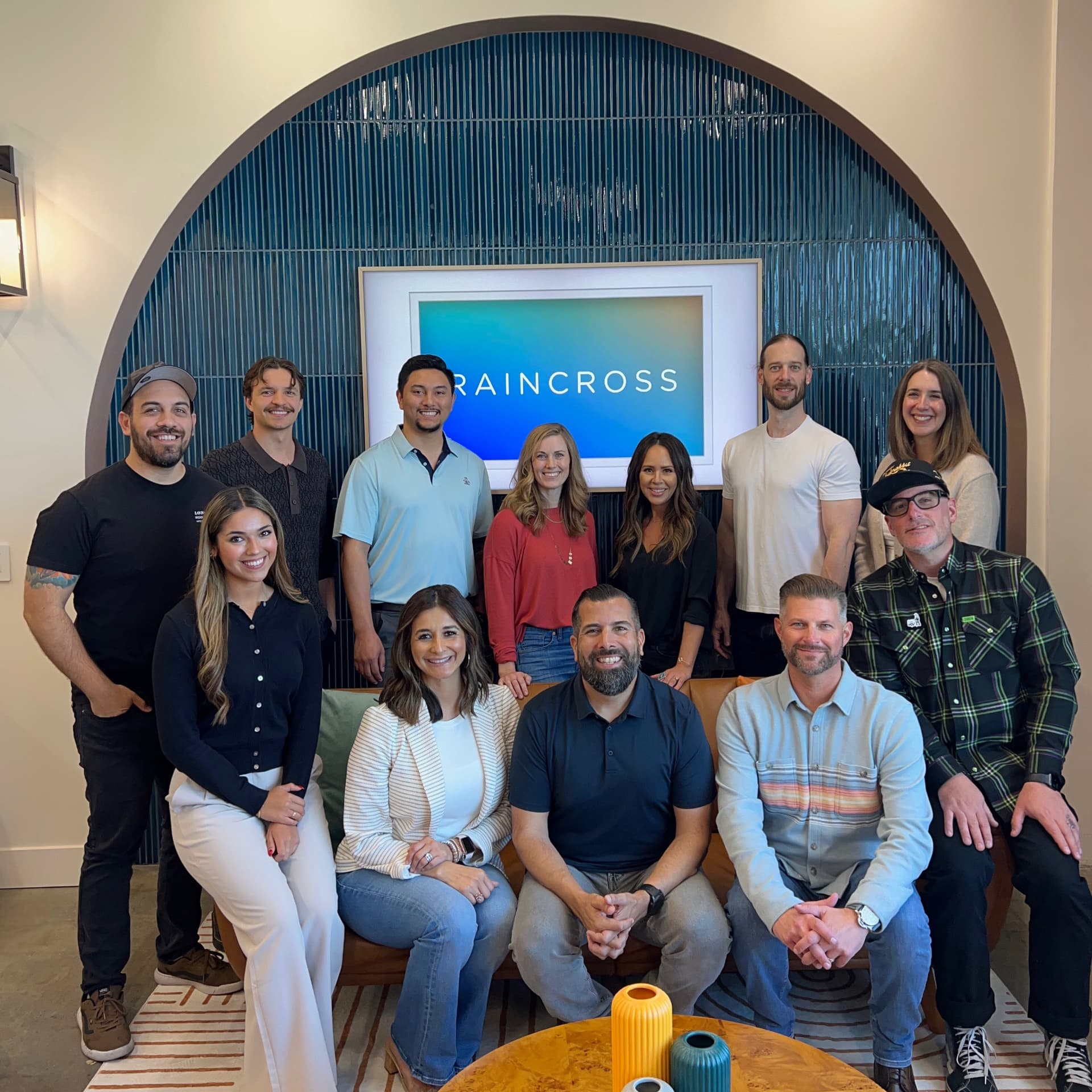 The Raincross team photographed together in the agency office, some seated and some standing, in front of an arched tiled wall and a screen showing the Raincross logo